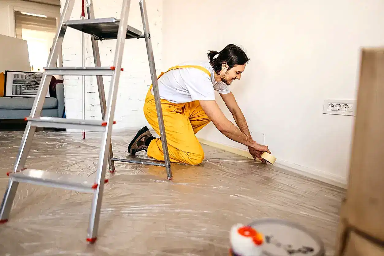 Boya badana işlemi öncesinde süpürgeliklere maskeleme bandı uygulayarak boya hazırlığı yapan sarı tulumlu profesyonel usta